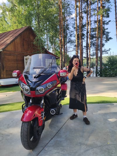 A woman with dark hair and sunglasses is standing next to a red and black motorcycle. She is wearing a black dress with a mesh overlay and a decorative skirt with fringe. She is holding a smartphone in her right hand and appears to be taking a selfie. The motorcycle is a Honda, with a large windshield. Behind the motorcycle is a wooden building with a red roof, and tall pine trees. The ground is paved with concrete and there is a patch of green artificial grass to the left of the motorcycle. In the background, there is a body of water, likely the Kama River, and a treeline. The scene appears to be in a park or recreational area in Chaykovsky, Russia.