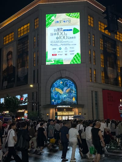 The image captures a lively street scene at night in Shanghai, China. The foreground is dominated by a crowd of people, suggesting a bustling urban environment and possibly an event or shopping activity. Many individuals are dressed in casual attire, and some are holding umbrellas, indicating that it might be raining or there's a threat of rain. The atmosphere appears to be one of general activity and movement.
Behind the crowd, a large building is prominently featured, adorned with illuminated signage and advertisements. One prominent display is a large LED screen showcasing a promotion for a "Boat Festival" with an offer for a "100 Yuan Consumption Voucher" and a "Membership Dining" event. Other signs advertise "SKINCEUTICALS" products, specifically an "AGE Cream" promising to reduce wrinkles and tighten contours, with a "30%" discount. Another large banner reads "你好, 新世界, 新丸中心!" which translates to "Hello, New World, New Wanhua Center!" identifying the location as the New Wanhua Center.
The architectural style of the building suggests a modern commercial establishment, likely a department store or shopping mall. The lighting from the signs and windows creates a vibrant and dynamic night-time ambiance. The presence of traffic lights and street lamps further reinforces the urban setting. The overall impression is that of a busy commercial district in Shanghai, drawing in shoppers and passersby with various promotions and attractions.