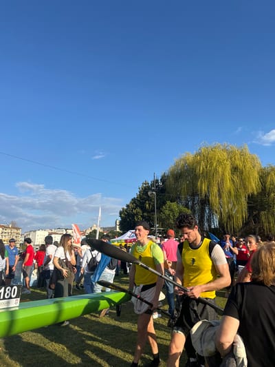The image depicts an outdoor sporting event or gathering in Mirandela, Portugal, under a clear blue sky with sparse clouds. The scene is primarily composed of a grassy area populated by numerous individuals.
In the foreground, two young adult males stand prominently, both wearing yellow sleeveless athletic tops and dark shorts. The man on the left, wearing a black baseball cap, holds a black and yellow canoe paddle and looks forward with a slight smile. To his right, the second man, with dark curly hair, holds a similar paddle and looks down, seemingly examining his equipment. A bright green canoe or kayak hull rests on the grass to the left of these individuals.
The midground features a dispersed crowd of people in various casual attire, standing and interacting. Partial views of white and red flags or banners are visible, along with parts of white tents, suggesting event infrastructure. A white sign with the number "180" and the text "JOGOS" is partially visible on the far left.
In the background, a residential area with multi-story buildings is situated on a gentle incline. A distinctive light-colored building with a dome or spire is visible on the distant horizon. A dense line of trees extends horizontally across the middle ground, with a large weeping willow tree, characterized by its bright yellow-green drooping foliage, dominating the right side of the frame. The overall lighting suggests a sunny daytime setting.