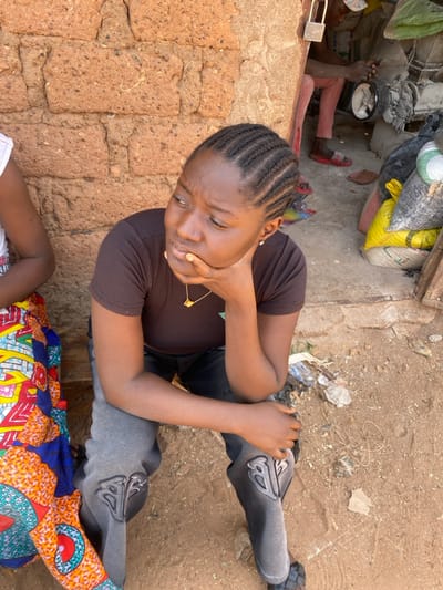 The scene is set outdoors in Rido, Nigeria, against a reddish-brown mud brick wall. In the foreground, a young Black female is seated, facing left, with her chin resting on her right hand. She has dark braided hair, wears a dark brown ribbed short-sleeved top, grey sweatpants with a prominent 'B' logo on the knees, a gold-colored necklace, and black woven slide sandals. To her left, another person is partially visible, wearing a colorful, patterned fabric. To the right, through a doorway, a male figure wearing a cap, light-colored pants, and red slide sandals operates a piece of machinery, possibly a grinding machine. Black and yellow sacks are stacked nearby, and a brass padlock hangs from the doorframe. The ground is unpaved reddish-brown earth with scattered debris.