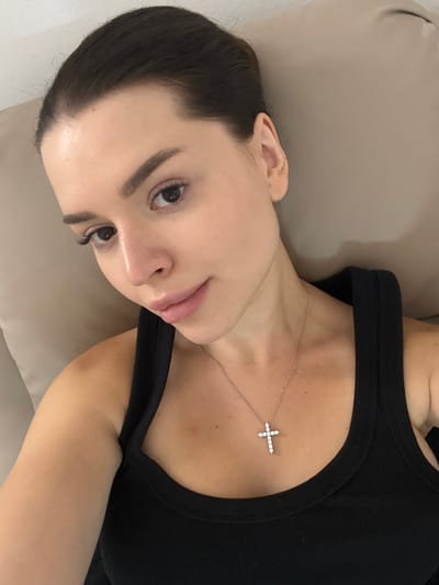 A person is lying down on a light brown surface, possibly a couch or bed. They are wearing a black tank top and a delicate silver necklace with a cross pendant. The cross is adorned with small, clear stones, likely diamonds. The person's hair is dark and pulled back, with some strands framing their face. Their face is turned towards the camera, with their left eye visible. They have full lips and dark eyebrows. The lighting suggests it might be indoors, with soft lighting. The orientation of the image is a rotation of 90 degrees counterclockwise from a standard upright view. The background is a neutral tone, creating a simple and focused composition on the subject. No visible text is present in the image. The context of Prague, Czechia does not provide any specific visual cues within the image itself.