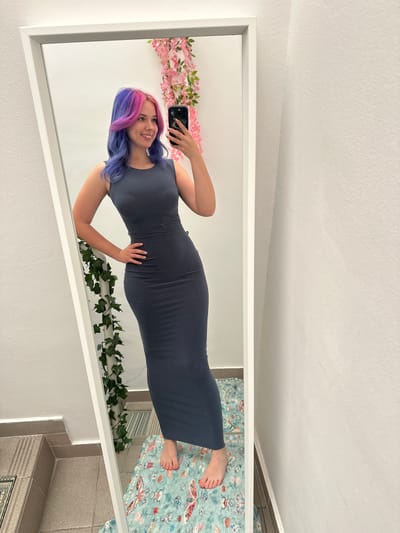 A young woman is captured in a full-body mirror selfie, standing indoors in what appears to be a residential setting in the City of Zagreb, Croatia. She is smiling broadly, looking directly into the smartphone she holds in her raised left hand. Her long, wavy hair is dyed in striking dual tones of vibrant pink and blue/purple. She is wearing a dark, form-fitting, sleeveless maxi dress, and her right hand is gently placed on her midsection. Her nails appear to be painted a light color.
Within the mirror's reflection, the backdrop behind the woman is decorated: a string of pink artificial cherry blossoms or similar flowers is visible on the left side, and a garland of artificial green ivy is draped along the bottom right edge. Below her bare feet, a light blue-green patterned rug or mat with illustrations of various insects (such as moths and beetles) and botanical elements is visible.
Outside the mirror's frame, the immediate surroundings of the room are partially visible. Above the mirror, a white, textured ceiling is seen. Below the mirror, the floor is covered in light gray tiles, with some sections featuring horizontal ridged patterns. The lighting suggests it is daytime. No discernible text is visible in the image.