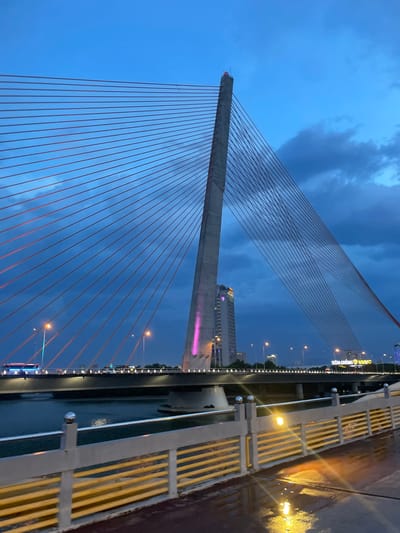 The image depicts a cable-stayed bridge at dusk, with a dramatic sky filled with dark clouds. The bridge's concrete pylon and numerous red cables are prominent subjects, stretching across the frame. In the background, city lights twinkle, and a tall building with pink and purple lights is visible. A yellow-railed pedestrian walkway runs along the right side of the bridge, with wet pavement suggesting recent rain. Streetlights illuminate the walkway, casting reflections on the wet surface. The overall mood is serene and slightly melancholic, enhanced by the twilight setting and the vast expanse of the sky. Visible text on a building in the background reads "TIỀU CƯỜNG VNVC". The time of day is clearly evening or dusk, with the sky transitioning from day to night. The location is identified as Đà Nẵng, Vietnam. No people are clearly visible in the image.