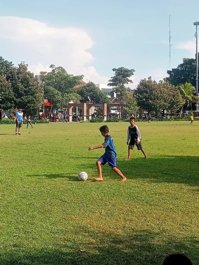 The image shows a green grassy field with three young boys playing soccer. One boy in a blue shirt is kicking the soccer ball with his right foot. Another boy in a black shirt is standing with his feet shoulder width apart. The third boy is walking away from the scene with a soccer ball at his feet. The field is located in the city of Pati, Indonesia, at latitude -6.753 and longitude 111.04. There are trees and a structure in the background. The sky is blue with white clouds. The sun is shining brightly. The grass is green and the light is strong. The boys are barefoot. The soccer ball is white. The structure in the background is made of brick.