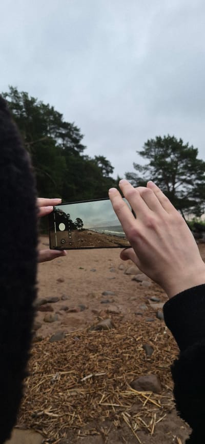 A person, viewed from behind, is holding a smartphone horizontally to take a picture of a beach scene. The phone screen displays a landscape with a shoreline, trees in the background, and a cloudy sky. The person is wearing a dark, fuzzy jacket. The foreground shows a sandy beach scattered with dry grass and rocks. The background features a dense line of trees on a gentle slope and the cloudy sky. The overall impression is of an overcast day at a coastal location. The image was taken in Repino, Russia.