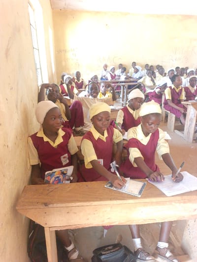 This image captures a crowded classroom in Mada, Nigeria, populated by numerous primary or middle school-aged students. The room features plain, light-colored walls with faint writing including "ROADS," "FRANCO," and "FRANCE," and is illuminated by natural light from a window on the left. Students are seated at rudimentary wooden desks and benches. In the foreground, three female students are visible. The two on the right are actively writing in notebooks with pens, while the leftmost student holds an open book and looks to her right. All three wear yellow short-sleeved shirts, maroon pinafores, and head coverings (two white, one yellow). In the midground, additional students, predominantly in similar maroon and yellow uniforms, are present. Several midground individuals are looking towards the camera, with two distinct students, one male and one female, making peace signs with their hands and smiling. School bags are visible beneath the desks in the foreground.