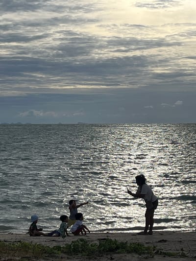 A silhouette of a woman taking a picture of children on the beach in Ko Samui District, Thailand. The children are sitting and standing on the sand near the water. The sky is cloudy and the sun is reflecting on the ocean. The scene is captured at dusk or dawn, with the sun low on the horizon.