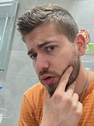 The image shows a close-up, indoor, selfie-style portrait of a young Caucasian male. He has light brown hair styled upwards and short stubble on his chin and jawline. His eyes are open and looking directly at the camera with a concerned expression. His right hand is raised, with his index finger and thumb touching his beard on his right cheek. He is wearing an orange t-shirt with thin black horizontal stripes and a silver chain necklace. The background consists of a tiled wall, a mirror on the left, and a shelf with a product featuring a woman's face on the right. The scene appears to be a bathroom, likely in a residential setting within the city of Yagodovo, Bulgaria.