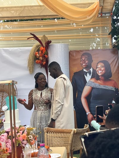 The scene depicts a celebratory event, likely a wedding or engagement reception, held under a draped cream-colored canopy in Kumasi, Ghana. In the central foreground, a dark-skinned woman with dark hair, wearing a silver sequined and beaded gown, smiles and holds a small red object in her left hand while gesturing towards a dark-skinned man standing to her right. The man, with short dark hair, wears a light beige long-sleeved tunic and observes the woman.
Behind the two individuals, a white fabric backdrop is adorned on the left with a large floral arrangement featuring gold, orange, and maroon elements. To the right, a prominent banner displays a photographic portrait of a smiling man in a black suit with a bow tie and a smiling woman in a black off-shoulder dress with white embellishments. To the far left, a multi-tiered cake with turquoise and white layers is visible on a gold-framed stand with wicker details.
In the immediate foreground, a table with a peach tablecloth holds a large floral arrangement of pink, orange, and white flowers, along with glassware and bottles, including a pink plastic bottle and a clear jar. Partially visible on the right, guests are present, with one individual in the extreme foreground holding a black smartphone vertically, seemingly recording or photographing the event. Wicker-backed chairs are visible at the table. The lighting suggests an outdoor or semi-outdoor setting during daylight hours.