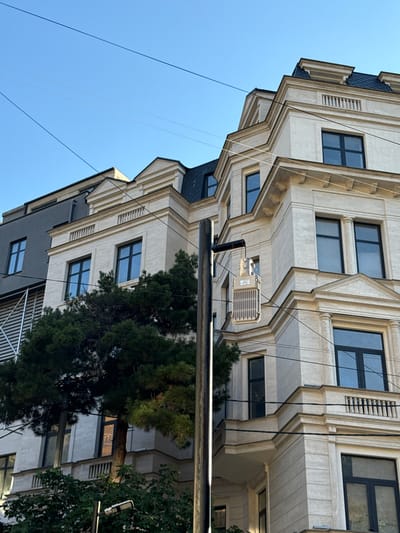 The image shows a view of a building in Tbilisi, Georgia, with a bright blue sky overhead. The building is multi-story and features light-colored stone or stucco with ornate architectural details, including cornices and decorative railings. Several windows with dark frames are visible. A large, dark green evergreen tree is partially obscuring the lower floors of the building on the left side. Overhead, several thin wires crisscross the sky, indicating the presence of utility lines. A dark pole with a gray, rectangular equipment box attached to it stands in the foreground, slightly off-center to the right. This equipment box appears to be an electrical transformer or a similar piece of infrastructure. The overall impression is an urban streetscape with a blend of older and modern architectural styles.