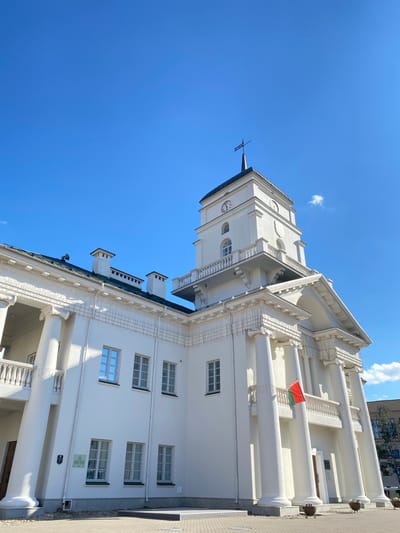 This is a tilted, upward-facing shot of a white neoclassical building with a tall clock tower under a bright blue sky. The building features classical architectural elements such as large columns, decorative cornices, and balustrades. A small flag, red and green, is visible on a balcony. The clock on the tower face shows the time as approximately 10:10. The scene appears to be outdoors on a sunny day, indicated by the clear blue sky and the bright lighting on the building. The architecture suggests a public building, possibly a government or historical landmark. The orientation of the photo, taken from below and tilted, emphasizes the height and grandeur of the structure. There are no people clearly visible in the frame. The location is indicated as Minsk, Belarus.