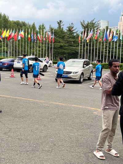 The image depicts an outdoor scene in Changchun City, China, likely on a college campus or at an event venue, given the presence of numerous national flags on poles lining a treeline. In the foreground, a person wearing a light brown t-shirt and checkered pants with white slides stands looking to the right. Behind them, three young men in blue athletic shirts and shorts are walking. The man on the left wears the number 10, the middle man wears number 11, and the man on the right wears number 1. Two cars are parked in the background: a silver Volvo SUV with a license plate reading "JA 2HP76" and a blue sedan. Orange traffic cones are placed on the asphalt. The sky is overcast.