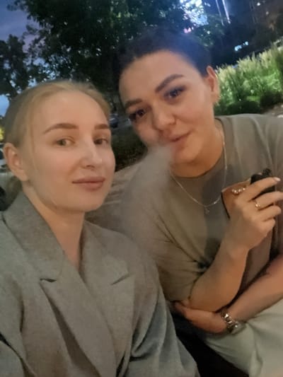 The media file is a close-up, low-angle selfie featuring two individuals. The person in the foreground, closer to the camera, appears to be a young woman with fair skin, blonde hair, and light-colored eyes. She is looking directly at the camera with a neutral expression. The second person, slightly further back and to the left, is a woman with darker hair and appears to be looking towards the camera as well. Her face is partially obscured by the first woman. Both individuals are wearing gray clothing, likely hoodies or t-shirts.

In the background, there are hints of a nighttime or evening setting. Out-of-focus greenery, possibly trees or bushes, is visible, with some areas illuminated by artificial light, suggesting an urban or park environment. The overall lighting is dim, consistent with dusk or nighttime. There are no prominent objects or text visible in the image, other than the jewelry worn by the person in the background, which includes a ring and a necklace. The image is slightly blurry, indicating it might have been taken in low light conditions or with quick movement. The emotion conveyed is casual and intimate, typical of a selfie taken between friends or partners. The context provided, Tyumen, Russia, does not offer specific visual cues within the image itself.