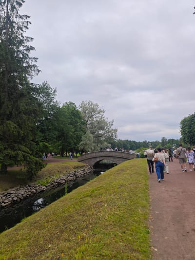 The image depicts a tranquil scene in the Catherine Park, city of Pushkin, Russia. A group of people are strolling along a dirt path that runs parallel to a narrow canal. The canal is lined with large stones and reflects the overcast sky. Across the canal, a stone bridge arches over the water, with more people visible walking on it. Lush green trees and foliage surround the path and canal, creating a serene park atmosphere. The sky is cloudy, suggesting a mild, possibly humid day.