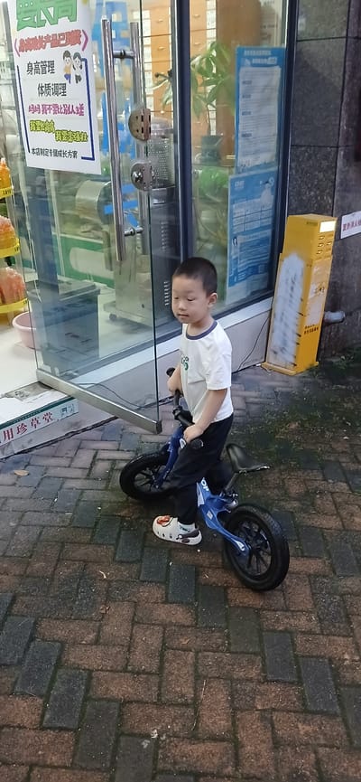 A young East Asian boy is standing on a blue balance bike on a wet, brick-patterned sidewalk in front of a glass storefront. The boy is wearing a white t-shirt with a blue collar, black pants, and white patterned clogs. He is looking to his right with a neutral expression. The storefront has glass doors with metal handles and a sign with Chinese characters. To the right of the storefront is a yellow fire alarm box. The background shows the interior of the store with shelves and plants visible through the glass. The scene appears to be in an urban environment, likely in China, as suggested by the Chinese text on the sign.