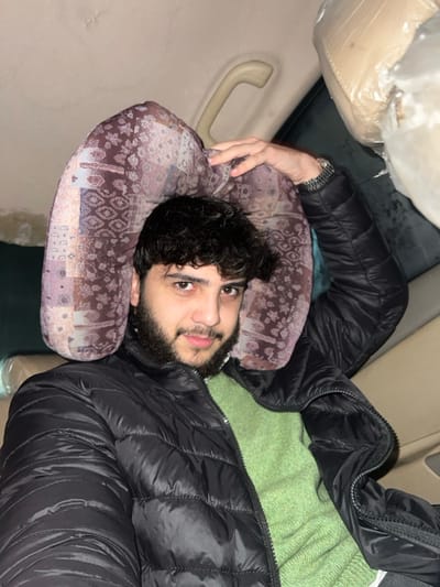 A man with dark curly hair and a beard lies on his side, using a patterned pillow. He wears a black puffer jacket. A green blanket is partially visible. The scene appears to be inside a vehicle, based on the interior elements like a plastic-covered seat and a grab handle. The overall perspective is slightly tilted.