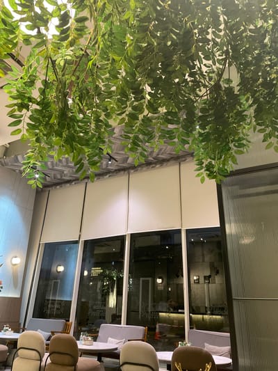 The image depicts the interior of a cafe or restaurant with a contemporary design, featuring suspended artificial greenery hanging from the ceiling. The space has large windows that look out onto a street scene. The interior includes modern furniture such as plush sofas and chairs arranged around tables. The lighting appears to be ambient, with some visible light fixtures on the ceiling and wall. Through the windows, reflections of the interior and the exterior can be seen, including the lights of what appears to be an urban environment. The overall impression is of a stylish and comfortable establishment, likely situated in a commercial or urban district. Without further contextual information or identifying markers within the image, it is impossible to definitively confirm the location in Central Jakarta, Indonesia.