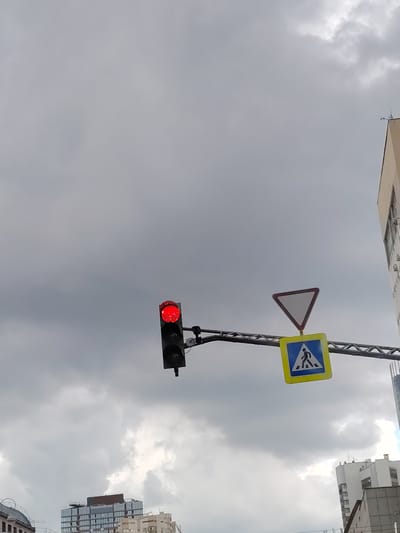 An outdoor, low-angle, vertical shot shows a traffic light with a red signal illuminated, a yield sign, and a pedestrian crossing sign against a cloudy, gray sky. The traffic light is on the left side of the frame. The yield sign, an inverted white triangle with a red border, is positioned to the right and slightly above the pedestrian crossing sign. The pedestrian crossing sign is a yellow square with a blue inner square, displaying a white silhouette of a person walking. The signs are mounted on a horizontal metal beam. In the background, the tops of several multi-story buildings are visible, including a modern glass-fronted building on the right, with lighter colored buildings below and to the left. The overall scene suggests an urban environment with overcast weather. This scene is located in Yekaterinburg, Russia.
