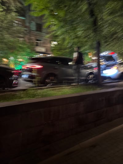 A blurry, long-exposure photograph taken at night in Yerevan, Armenia. The photograph shows a street scene with trees and buildings lining one side of the road, and a concrete wall or structure on the other side. Motion blur is evident throughout, indicating camera movement during the exposure. Several cars with headlights and tail lights are visible on the road. Buildings are also visible, although blurred. Vegetation is present, primarily trees along the street. The overall environment suggests an urban setting, and it is likely taken at night due to the presence of artificial lights and the darkness of the background.