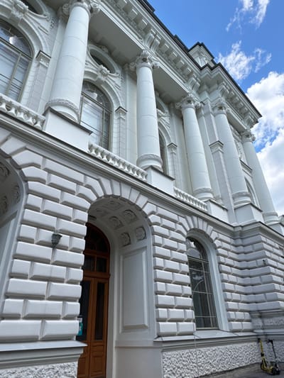 The image shows the facade of a white building with classical architectural elements, including large columns and arched windows. The building has a textured white stone exterior. In the foreground on the right, there are two electric scooters parked on the sidewalk. The sky above is blue with scattered white clouds. The image appears to be taken from a low angle looking up at the building.