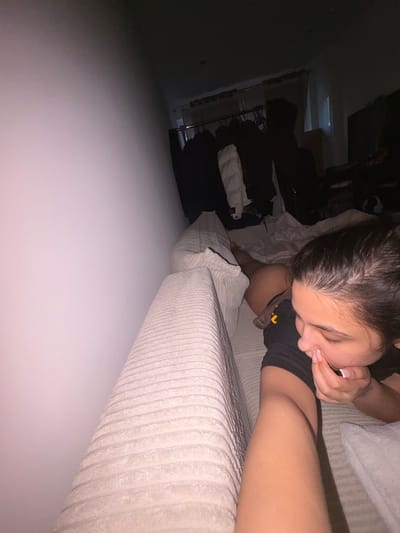 A close-up, eye-level shot captures a young person lying on a textured couch in what appears to be a bedroom. The person, with dark hair and wearing a black shirt, is looking down and touching their face with their fingers, particularly their nose. Their gaze is directed towards their own leg, which is extended in front of them and partially visible. The surrounding environment is dimly lit, with a wall on the left side of the frame. In the background, clothing items are hanging on a rack, suggesting a closet area. The overall impression is that of a private, intimate moment. This scene is taking place within a private residence, an apartment or house, in Mönchengladbach, Germany.