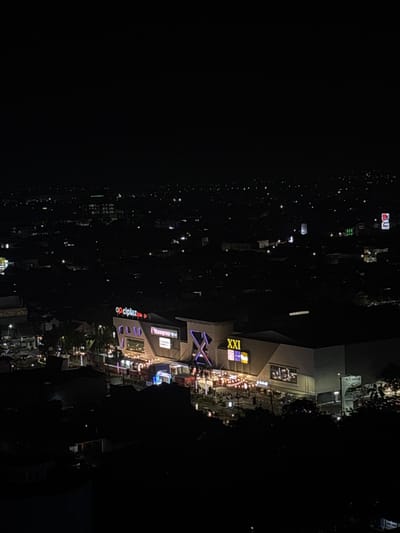 An aerial nighttime view depicts a brightly illuminated commercial complex, identified by prominent neon signs for "ciplaz," "Transmart," and "XXI" cinema, situated within the city of Sidoarjo, Indonesia. The complex features modern, angular architecture with multiple levels. An outdoor area directly in front of the main entrance is lit by numerous string lights, beneath which a gathering of individuals is observed. Vehicle lights are visible on the street immediately adjacent to the complex. The surrounding environment is an urban landscape, extending into the dark background as a sprawling network of scattered residential and commercial lights beneath a clear, starless sky. No specific interactions are discernable, but the presence of many people indicates public activity.