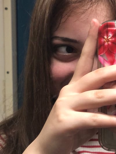 The image depicts a close-up, cropped portrait of a female subject located in the city of Saverne, France. The subject's long, dark brown hair frames the visible right side of her face. Her right eye is open and directed towards the viewer's left. Her right hand is prominent, held up in front of her face, with fingers partially obscuring the central facial features. This hand holds a smartphone vertically, encased in a transparent or clear protective cover featuring a red floral design with visible yellow stamens. A portion of the subject's clothing, characterized by red and white horizontal stripes, is visible below her hand. The background is out of focus, revealing a light-colored wall or surface with two small dark circular elements on the left, next to a vertical blue-colored object or surface. The composition suggests a mirror selfie.