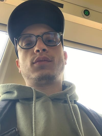 A person is depicted in a close-up, upward-facing selfie or portrait shot, rotated 90 degrees counter-clockwise. The individual, appearing to be male, is seen from the chest up, looking directly towards the camera with a neutral expression. They are wearing a dark, possibly black, baseball cap pulled low over their forehead, and dark-rimmed glasses. The person has facial hair, appearing as stubble or a short beard, around their chin and cheeks.

They are dressed in a light green or olive green hooded sweatshirt, with the hood visible around the neck and drawstrings hanging down the front. A black strap, likely from a backpack or bag, is visible over their right shoulder (left side of the image as displayed).

The background suggests an interior setting, possibly public transportation like a train, tram, or bus. A bright, overexposed area on the right side of the image indicates a window or skylight, letting in a significant amount of daylight. On the upper left side of the image (which would be the ceiling or upper panel if the image were correctly oriented), a circular, light-colored button with a green center is visible. Within the green center, there is a stylized symbol that appears to be "BX" or "XB". The surrounding interior panels are light-colored.