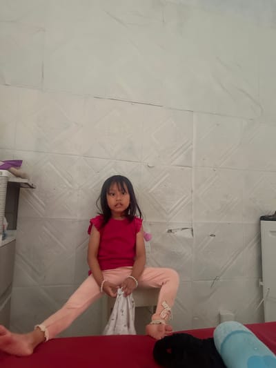 The image shows a young girl with dark hair and bangs, wearing a pink shirt and pink pants. She is lying on her side on a red surface, with her legs bent. Her pants have decorative bows on the ankles. She is looking at the camera with a slight smile. Her right hand is holding onto a white cloth. Beside her is a light blue cylindrical object. The background appears to be a wall with a patterned texture. The scene is likely indoors, given the presence of the red surface and the wall texture. The lighting suggests it is daytime. No text is visible in the image. The location context provided is Kecamatan Pati, Indonesia.