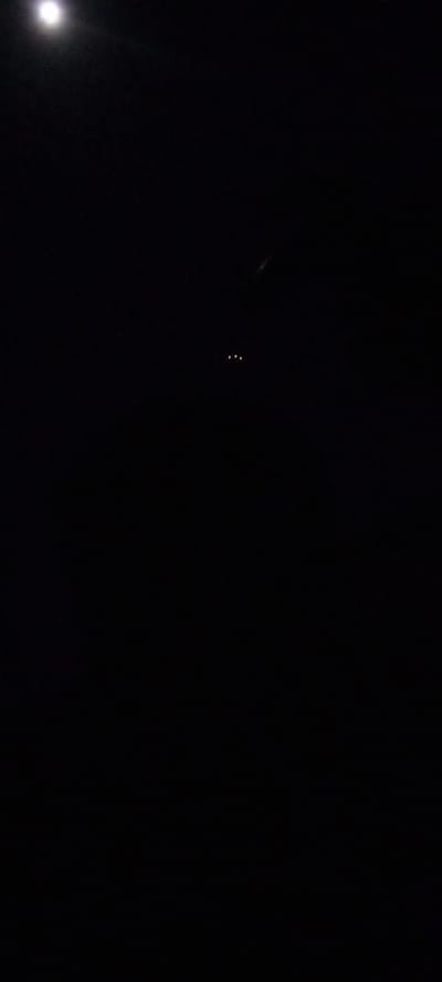 The image presents a predominantly dark, nocturnal scene, featuring minimal light sources against a black background. In the upper-left quadrant, a bright, diffused white light is visible, likely representing the moon due to its intensity and halo effect, though it appears overexposed. Towards the center-right, three small, distinct, yellow-orange lights are horizontally aligned, suggesting artificial illumination from a distant source such as a vehicle or structure. Faint, elongated streaks of light are subtly visible in other dark areas, possibly indicating reflections or very distant light trails. No other discernible objects, people, or activities are present. The pervasive darkness clearly indicates a nighttime setting. There are no specific visual cues within the frame to confirm the location as Ilogbo Eremi, Nigeria, nor is any text visible.