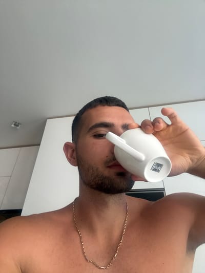 A man with short dark hair and a beard is holding a white mug to his lips, taking a drink. He is shirtless and wearing a gold chain necklace. The background is a modern kitchen with white cabinets. The image is a selfie taken from a low angle. The location is not specified as Gabrovo, Bulgaria.