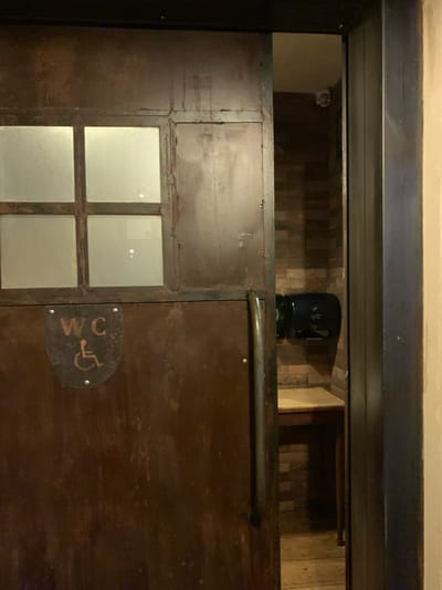 The image displays a dim, indoor doorway, likely an entrance to a public restroom, in Marousi, Greece. The central subject is a dark brown door, possibly made of wood or metal with a textured, distressed finish. On its left side, four square frosted glass panes are arranged in a grid. A long, curved metal handle is mounted horizontally on the right side of the door.
Below the handle, a circular, metallic sign is affixed, featuring the text "WC" and a universally recognized wheelchair accessibility symbol. The doorway itself is framed by a sturdy, dark-colored, possibly metal structure.
Above the door, an open space reveals a deeper interior. A light-colored wall with a subtle pattern, possibly wood or tile, is visible. On a ledge within this space, two objects are discernible: a small, light-colored device, possibly a security camera, on the far left, and a larger, dark, cylindrical object, potentially a speaker or another camera, positioned further right.
The ambient lighting is low, suggesting an indoor setting during the evening or night. No people are present, and no specific activity is depicted, presenting a static scene of the entrance.
