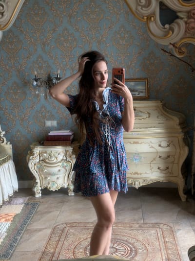 The image shows a young woman taking a mirror selfie in a lavishly decorated room. The room features ornate, antique-style furniture, including a cream-colored wardrobe with gold accents and intricate carvings. The walls are covered in blue wallpaper with a gold damask pattern. A large, decorative mirror with a golden frame is visible, reflecting the scene.
The woman is wearing a short, blue floral-print dress with a ruffled white collar. She is holding an orange smartphone in her right hand, angled to capture her reflection. Her left hand is touching her hair. She has long, dark brown hair and is looking at the phone's screen.
On a shelf next to the wardrobe, a stack of books is visible. There is also a small framed painting on the wall in the background. The floor is tiled, and a decorative rug with intricate patterns is partially visible. The overall impression is one of opulence and a vintage aesthetic. The lighting suggests it could be daytime, but there are no direct clues about the time or weather. The woman's expression is neutral, focusing on taking the selfie. There is no visible text in the image.