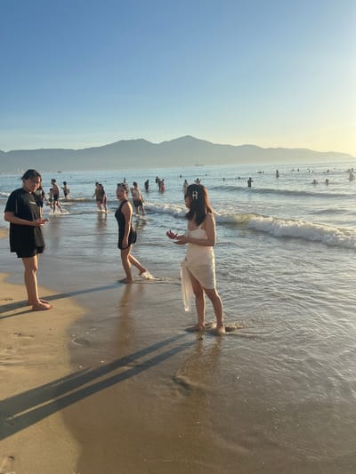 The image depicts a beach scene in Da Nang, Vietnam, with a clear sky and distant mountains. Several people are visible, some wading in the shallow ocean water and others on the sandy shore. In the foreground, two women are standing on the wet sand. The woman on the left, wearing a black t-shirt and shorts, is holding a mobile phone and looking at the woman to her right. The woman on the right, dressed in a white cover-up over a black swimsuit, is looking towards the ocean and has her hand near her mouth. Further out in the water, a crowd of people are swimming and standing. The sun appears to be setting or rising, casting long shadows on the sand and creating a warm, golden light across the scene. Waves are gently breaking on the shore, indicating a calm sea.