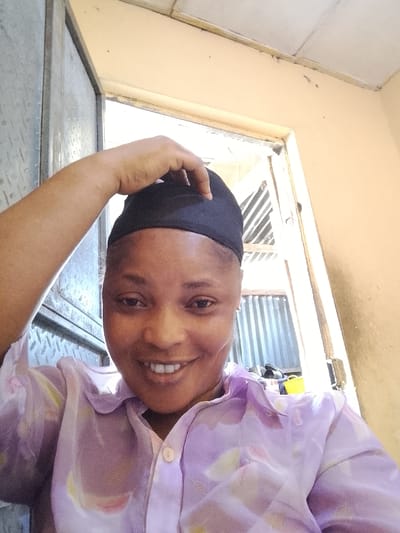 A smiling woman wearing a light purple, floral-patterned button-down shirt and a black head covering poses indoors. She is touching the top of her head covering with her right hand. Behind her is a doorway, showing a metal-corrugated structure. The background also includes an off-white wall and a partially visible window.