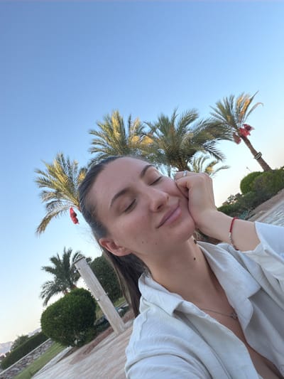 A young woman with her eyes closed leans her head against her hand in front of a clear blue sky with palm trees. She is wearing a white shirt. A red string bracelet is visible on her wrist. The scene appears to be in Sharm El Sheikh, Egypt, suggested by the palm trees and warm climate.