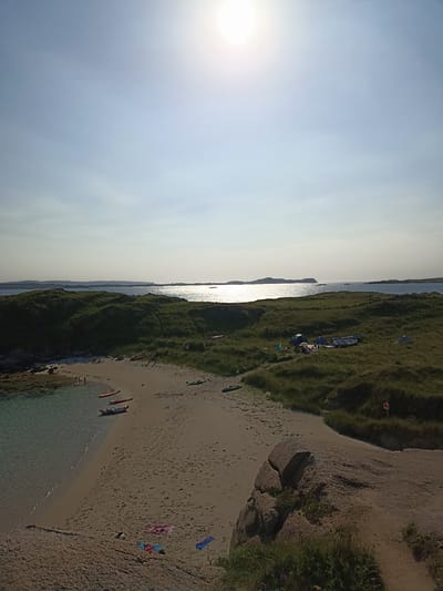 The image depicts a serene coastal landscape on a bright, sunny day. In the foreground, a sandy beach slopes down to clear, shallow water. Several kayaks are resting on the sand and in the water, suggesting recreational activity. Scattered towels add a touch of color to the beach.
Further inland, rolling green hills dotted with patches of grass and rocks dominate the scene. A small campsite is visible on one of the hills, featuring several tents and a small boat. In the distance, the calm, sun-drenched ocean stretches out, meeting the horizon where islands can be seen. The sun is high in the sky, casting a bright glare on the water.
The overall impression is one of a peaceful, natural environment, likely a secluded bay or cove, ideal for outdoor activities like camping and kayaking. The scene captures the beauty of a coastal summer day. The location is specified as Carnboy, Ireland.