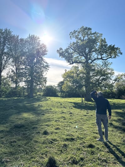 This image captures an outdoor scene on a bright, sunny day in a grassy field or park, characteristic of North Hertfordshire, United Kingdom. The frame, oriented correctly, features a clear blue sky with some scattered clouds, and a prominent sun flare in the upper left corner, indicating the sun's high position.
In the mid-ground on the right side, a man stands facing left, dressed in a dark long-sleeved top and lighter-colored trousers. He has his right hand raised to his forehead, appearing to shield his eyes from the direct sunlight as he looks forward or slightly upward.
Several large, mature deciduous trees with dense green foliage are scattered across the left and central parts of the landscape. Their branches, though partially obscured by bright sunlight in some areas, show intricate patterns. These trees cast long, distinct shadows across the vibrant green grass, suggesting either morning or late afternoon. The terrain is gently undulating, covered in lush green grass that shows variations in texture and height. The overall atmosphere is peaceful and serene, emphasizing the natural beauty of the sunlit outdoor environment. No text is visible in the image.