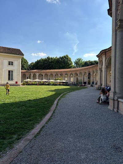 This image depicts an outdoor scene at the Villa Arconati-Far in Castellazzo di Bollate, part of the metropolitan area of Milan, Italy, specifically showcasing its distinctive D-shaped portico. The scene is illuminated by bright sunlight under a clear blue sky with scattered white clouds.
In the foreground, a dark grey gravel path curves from the bottom right towards the center, bordered on its left by a narrow strip of paving stones and a vibrant green lawn. A young boy, dressed in a yellow t-shirt and dark shorts, runs from left to right across this lawn, casting a distinct shadow.
To the left, a portion of a light-colored building with a reddish-brown tiled roof and rectangular windows is visible. A red box, possibly a fire alarm or emergency equipment, is mounted on its exterior wall. Dense green trees form a backdrop behind this building and continue across the midground.
The midground is dominated by a grand, curved colonnade, characteristic of the Villa Arconati's architecture. This structure features numerous arches supported by light-colored columns and pillars, topped with a reddish-brown tiled roof. Along the base of the colonnade, particularly on the left half, several white sun umbrellas are open, sheltering tables and chairs, suggesting an outdoor cafe or seating area. Potted plants or flower beds are arranged in front of this section.
To the right, closer to the viewer, the side of a large, light-colored column from a similar architectural structure is visible, framing the right edge of the image. Several individuals are seated on a bench or ledge in the shaded area beneath this structure. These include a woman in a white top and dark pants viewed from the back, and another person in a striped top beside her. Other individuals are seated further along the bench, relaxing and observing the scene.