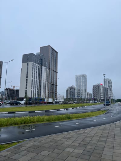 This image depicts a modern urban scene in Yekaterinburg, Russia. The primary subjects are several tall, contemporary apartment buildings with a mix of white, gray, and dark glass facades. These buildings are arranged along a wet, asphalt road, indicating recent rain. The road is lined with a strip of vibrant green grass and has white lane markings. A paved walkway made of square gray tiles runs alongside the road.
Cars are parked along the side of the road, suggesting a residential or commercial area. In the background, more buildings are visible, including some under construction, with a crane indicating ongoing development. Streetlights are present along the road. The sky is overcast and a uniform light blue-gray, suggesting a cloudy or possibly rainy day. The time of day appears to be daytime, but the overcast conditions create a somewhat subdued light.
There are no people clearly visible in the image, nor is there any discernible text. The overall impression is of a new, developing urban environment in Yekaterinburg, characterized by modern architecture and infrastructure, under a neutral, cloudy sky.