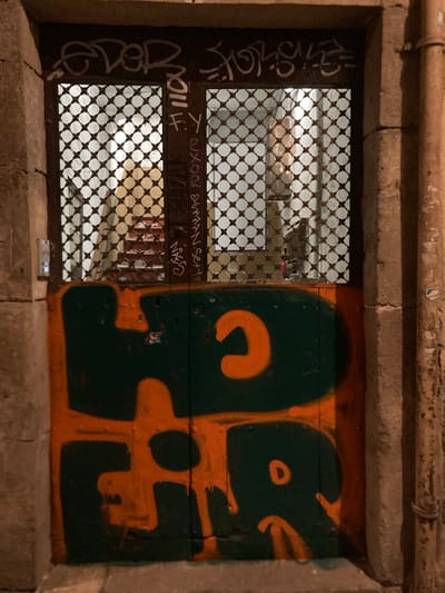 The image depicts an outdoor scene, likely in Barcelona, Spain, at night. It shows a recessed area in a stone wall, possibly a basement window or utility access point. The left side of the opening is covered by a metal grate, revealing a dimly lit interior with a checkered pattern, suggesting more grates or a patterned wall. White graffiti is visible on the metal frame of the grate and on the stone surround.

The right side of the opening is covered by a wooden panel that has been painted with bold, stylized graffiti in green and orange. The orange graffiti features large, abstract shapes that form letters or symbols. A small, square object, possibly a device or a piece of trash, rests on the stone ledge below the opening. The overall impression is one of an urban, somewhat neglected space marked by street art. There is no visible indication of the weather, and the emotional tone is neutral, focusing on the visual elements of the scene.

Visible text includes white graffiti on the left side which is difficult to decipher due to its stylized nature and low resolution. Some words that might be present are "CYOC," "H," "K," and potentially "OVER." On the wooden panel, the orange and green graffiti consists of abstract shapes that are difficult to read as clear text.