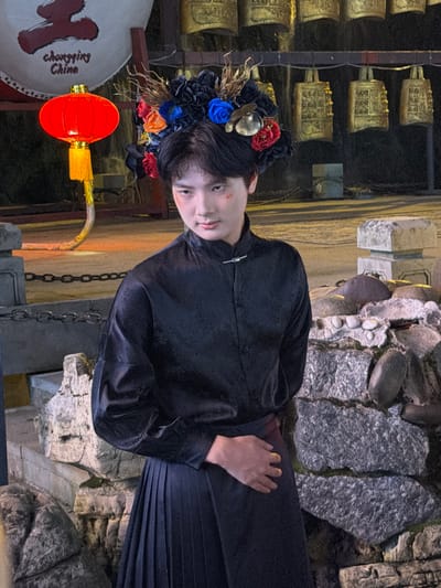 A person adorned with a floral headdress featuring blue, red, and orange flowers is captured in a portrait. They are wearing a dark, possibly black, traditional Chinese garment with ornate patterns and a pleated skirt. The person's face is turned slightly towards the viewer, with their gaze directed outwards, conveying a subtle expression.
The background suggests an outdoor setting with elements of traditional Chinese architecture and natural rock formations. Golden bells or chimes are visible on the left, arranged in rows, hinting at a cultural or historical site. To the right, rough-hewn rocks and moss create a textured backdrop. A red lantern with yellow accents hangs prominently in the foreground, adding a vibrant splash of color. The overall lighting indicates it might be evening or nighttime, with artificial lights illuminating the scene.
On a large white circular object, possibly a drum, text is visible: "Chongqing China" written in a calligraphic style. The presence of the lantern and the text explicitly places the scene in Chongqing, China. The image appears to be a posed portrait, possibly part of a cultural event or photoshoot celebrating traditional Chinese aesthetics.