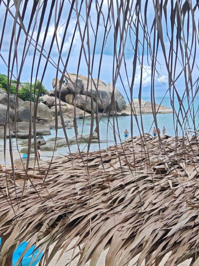 The image depicts a beachfront scene in Baan Chaweng Noi, Thailand, viewed through a thatched roof made of dried palm leaves. In the foreground, the dry leaves obscure much of the lower portion of the image. Beyond the leaves, the turquoise ocean meets a sandy beach dotted with large, weathered rocks. Several individuals are visible near the water's edge. Two men are standing in the shallow water, one wearing blue shorts and the other possibly a darker garment. Further out, a group of three people, two men and a woman in a bikini, are also in the water. Another person is partially visible further to the right. A blue kayak is positioned on the sand in the lower left corner of the image, partially obscured by the thatch. The sky above is a mix of blue with scattered clouds. Lush green vegetation is visible on the left side of the frame, behind the rocks.