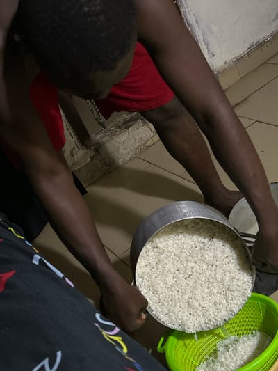 This media file shows a close-up, overhead view of two people and a large amount of uncooked white rice. The primary subject is a person wearing a red garment, whose dark-skinned legs and arms are visible. They are holding a metal pot filled with rice. To the right of this pot, a bright green plastic colander also contains rice. The scene appears to be indoors, possibly a kitchen or dining area, with tiled flooring visible. The lighting suggests it might be evening or a dimly lit indoor space. The overall mood is neutral, focusing on the task of handling the rice. There is no visible text in the image. The context provided places this scene in Ihiagwa, Nigeria.