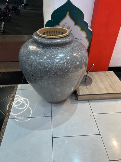 In Depok, Indonesia, a large, speckled gray vase with a tan plastic insert sits prominently on a tiled floor. To its right, a small metal stand emerges from a light brown wooden platform. Behind the vase, a red and white wall features a decorative green Islamic motif. In the background, motorcycles are parked, and a few people are visible, suggesting an outdoor or semi-outdoor public space, potentially near a mosque or a marketplace. The lighting indicates it is either evening or an indoor setting with artificial lights.