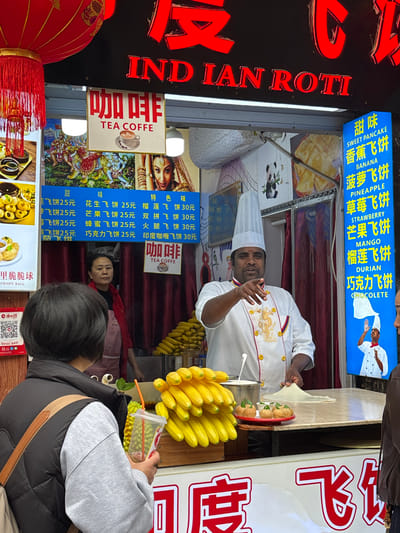This image depicts a street food stall in Shapingba District, China, specializing in Indian Roti.

At the center, a man identified as a chef, wearing a white chef's coat with red and gold trim, a tall white toque, and a dark complexion, stands behind a counter. He gestures with his right hand forward and slightly to the right, while his left hand rests on a circular piece of dough being prepared on the counter. To his left, a large stack of bright yellow bananas is displayed on the counter, next to a metal pot. Further left, a small red tray holds three small, round, possibly fried food items.

Behind the chef, another woman with dark hair, wearing a red top and a light-colored apron, stands observing. Behind her, a large quantity of corn cobs is stacked.

In the foreground, a person with short dark hair, viewed from the back, wears a grey long-sleeved shirt and a black quilted vest. This person holds a clear plastic cup with a straw, containing a beverage, and faces the food stall.

Above the stall, a large red neon sign displays "INDIAN ROTI" in English and corresponding Chinese characters. Below it, several smaller signs advertise "TEA COFFE" and various roti flavors in both Chinese and English (e.g., Banana, Pineapple, Strawberry, Durian, Chocolate), with prices listed in Yuan. Posters featuring images of Indian deities and a woman are also visible. A blue vertical sign on the far right reiterates roti flavors. Red decorative lanterns hang on the left side of the stall. A QR code for mobile payment is visible on the far left.