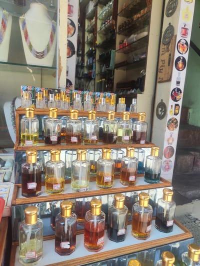 The image shows a display of perfume bottles, likely for sale, in Puttaparthi, India. The bottles are arranged in multiple rows and columns in a wooden-framed glass cabinet. Each bottle is rectangular with a gold-colored cap and a label indicating the perfume's name or scent. The perfumes come in various colors, including clear, yellow, amber, and dark brown. The bottles are labeled with names like "Royal Guquet," "Bakur," "BISCUIT," and "Gold Sandal". The shelf below the perfumes displays necklaces and other jewelry. Above the perfumes is a decorative panel featuring images of perfume bottles and a sign that reads "em Ply N". The location appears to be an outdoor market or shopfront in Puttaparthi, India. The scene suggests a retail environment with a focus on perfumes and jewelry.