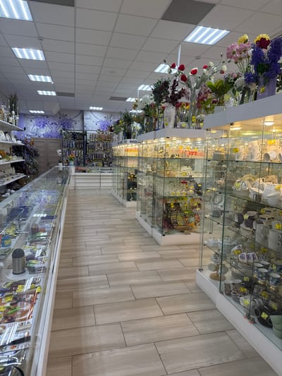 The image shows the interior of a shop in Klin, Russia. The shop appears to specialize in housewares, gifts, and decorative items. On the left, artificial flowers and foliage are displayed on shelves, and on the right, glass cabinets are filled with glassware, cups, plates, and other ceramic items. In the distance, there are displays of other merchandise, including what looks like gardening tools. The floor is tiled in light brown rectangular tiles, and the ceiling has recessed rectangular lighting panels. The overall impression is that of a well-lit and organized retail space.