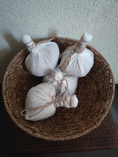 The image shows a woven basket filled with four white cloth bundles tied with twine. The bundles appear to be massage herbs or aromatherapy sachets. The basket is placed on a dark, woven mat, and the background is a textured, light-colored wall. The lighting suggests an indoor setting, possibly a spa or a home where these items are stored. The overall impression is one of calm and relaxation, with a focus on natural materials and wellness. There is no visible text in the image.