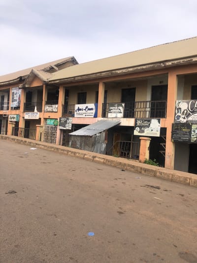 The image shows a street scene in Barakin Lerawi, Nigeria, with a two-story building housing multiple businesses lining the right side of the road. The building has an orange and cream facade and a beige metal roof. Several signs are visible, advertising businesses such as "SHUP'S AGRO VET", "CARTER PEF COLLECTION", "J&J CLOTHING ACCESSORIES", "Aico's Corner", and "De Fajo's EMPIRE". The street itself is paved and appears to be slightly sloped. There are some papers scattered on the ground near the curb. The sky is overcast.